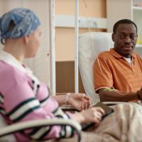 A black man and a white woman with a head scarf talking during chemotherapy treatment in clinic.